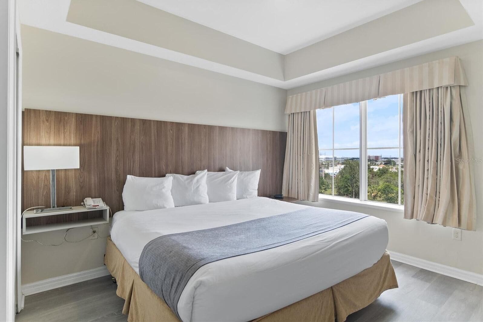 A hotel room with a large bed, a bedside table with a lamp, a view of the city through the window, and a wooden headboard.