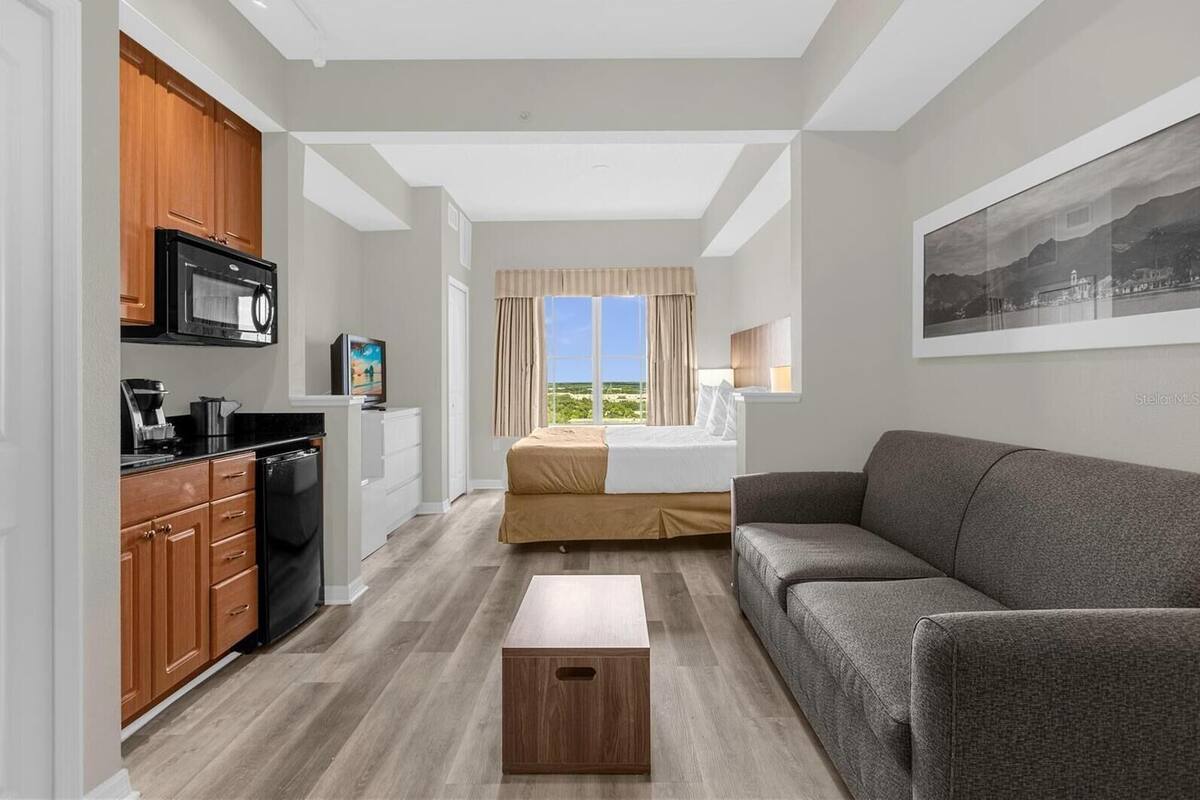 A modern hotel room with a kitchenette, a bed with a view, a grey sofa, and a wooden coffee table.