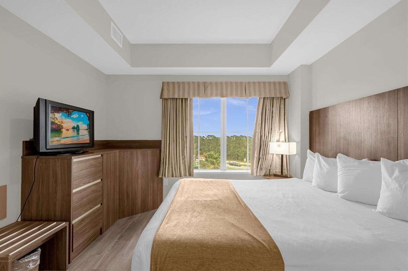A hotel room with a large bed, a TV on a wooden dresser, a window with curtains, and a view of the outdoors.