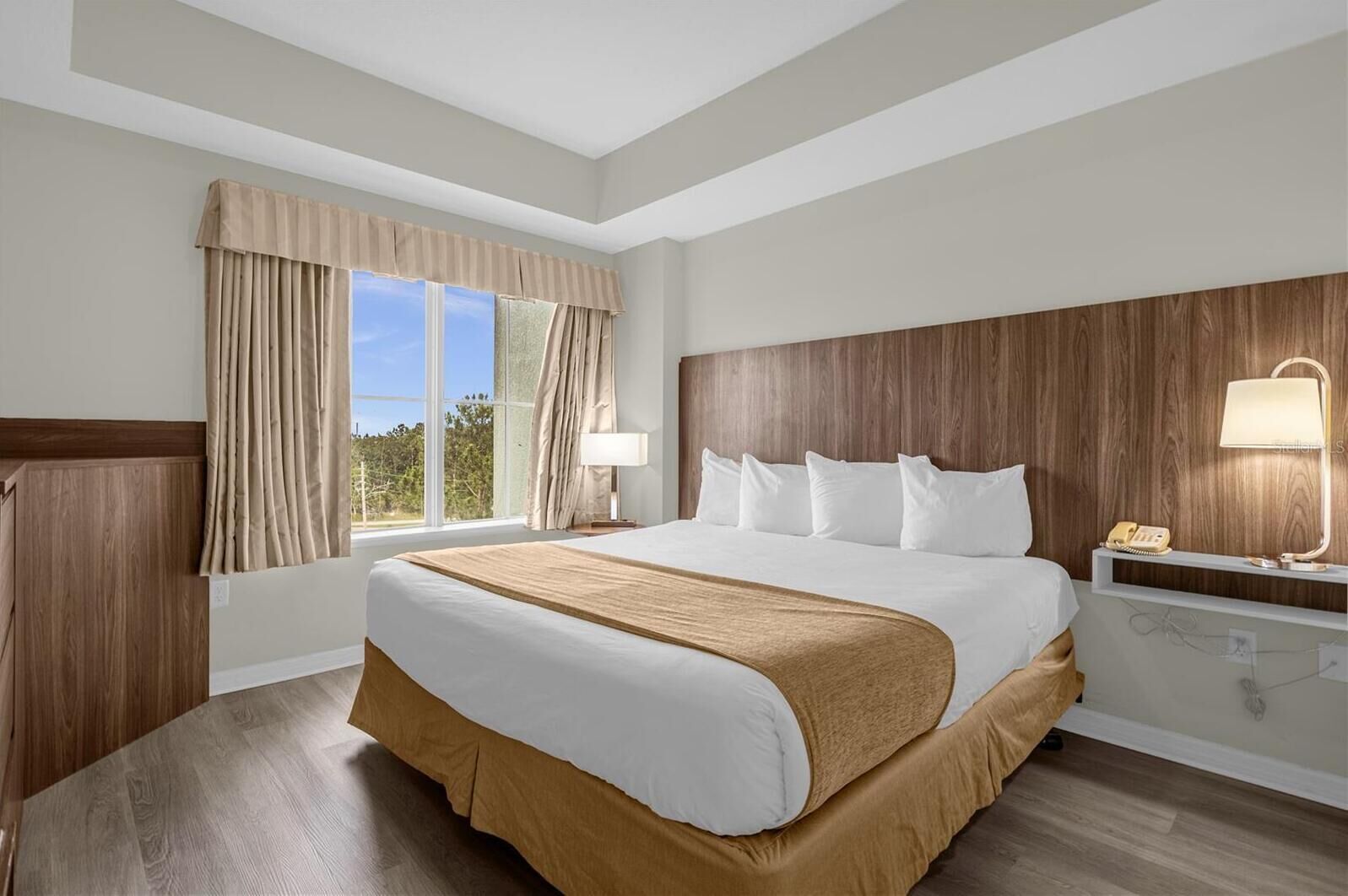 Habitación de hotel con una cama grande, cabecera de madera, una mesita de noche con lámpara y una ventana con cortinas.