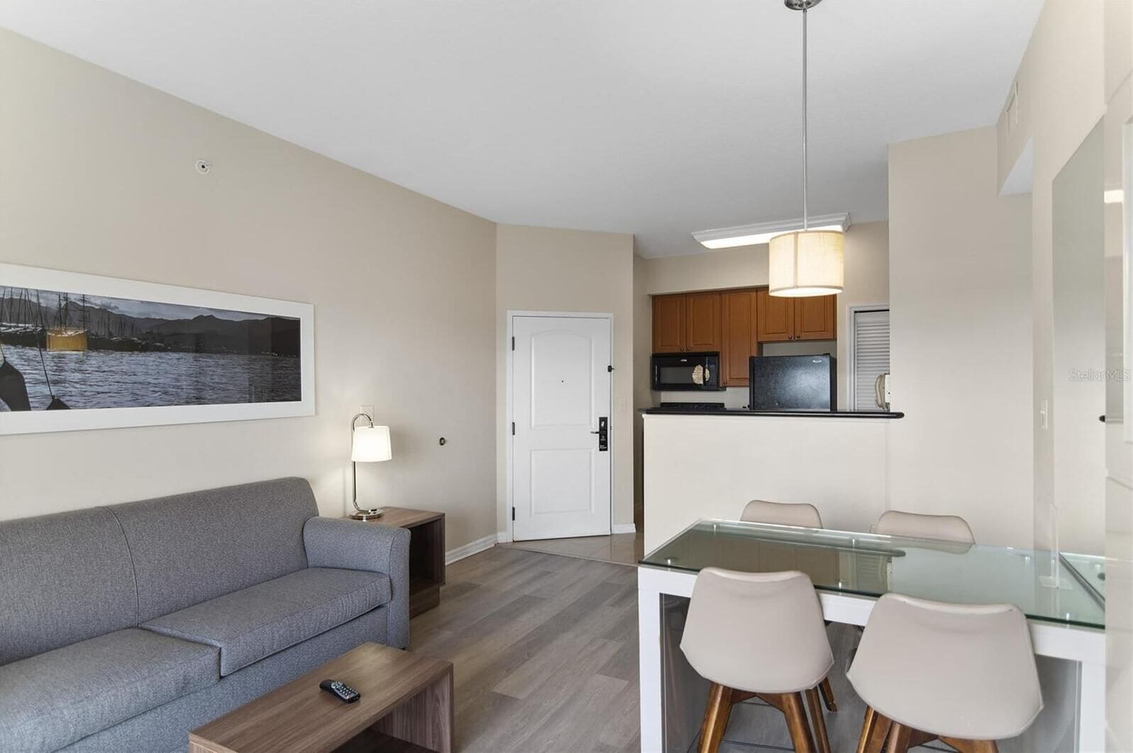 Una sala moderna con un sofá gris, una mesa de comedor de cristal con dos sillas, una mesita auxiliar con lámpara, y una zona de cocina con armarios de madera y microondas.