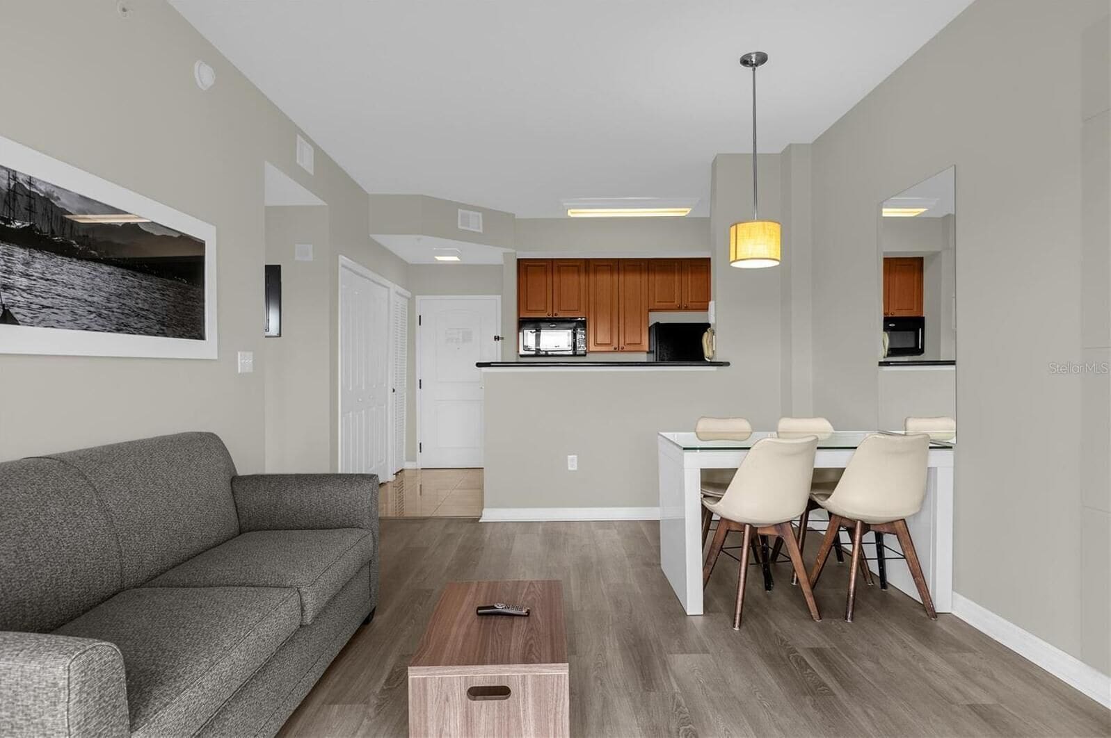 A modern living room with a gray sofa, a dining area with white chairs and a glass table, and a kitchen with wooden cabinets.