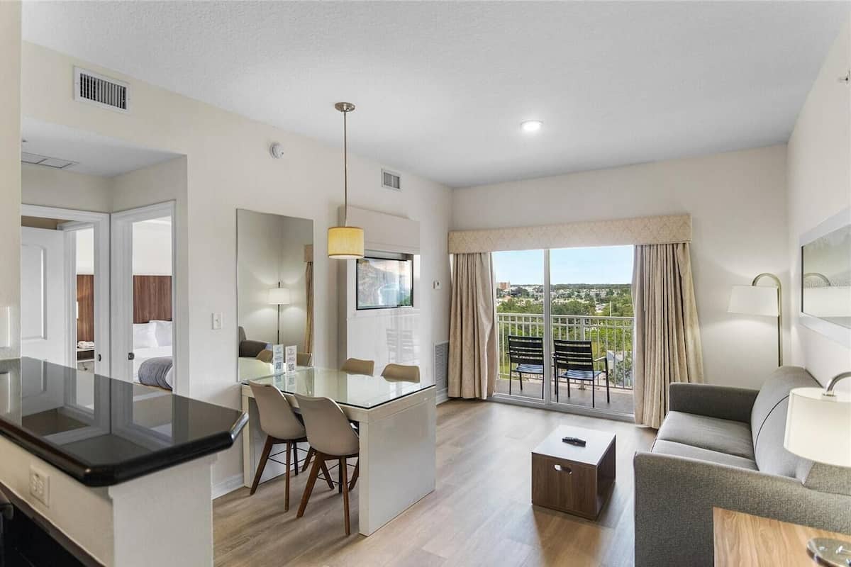 A modern hotel room with a dining area, a sofa, a balcony with curtains, and a large window.