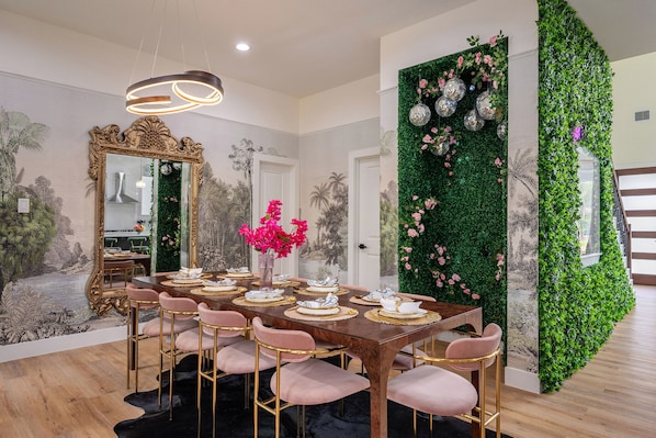 Un coin repas avec une table en bois dressée pour six personnes, entourée de chaises roses, d’un grand miroir décoratif et d’un mur vert agrémenté d’éléments décoratifs.