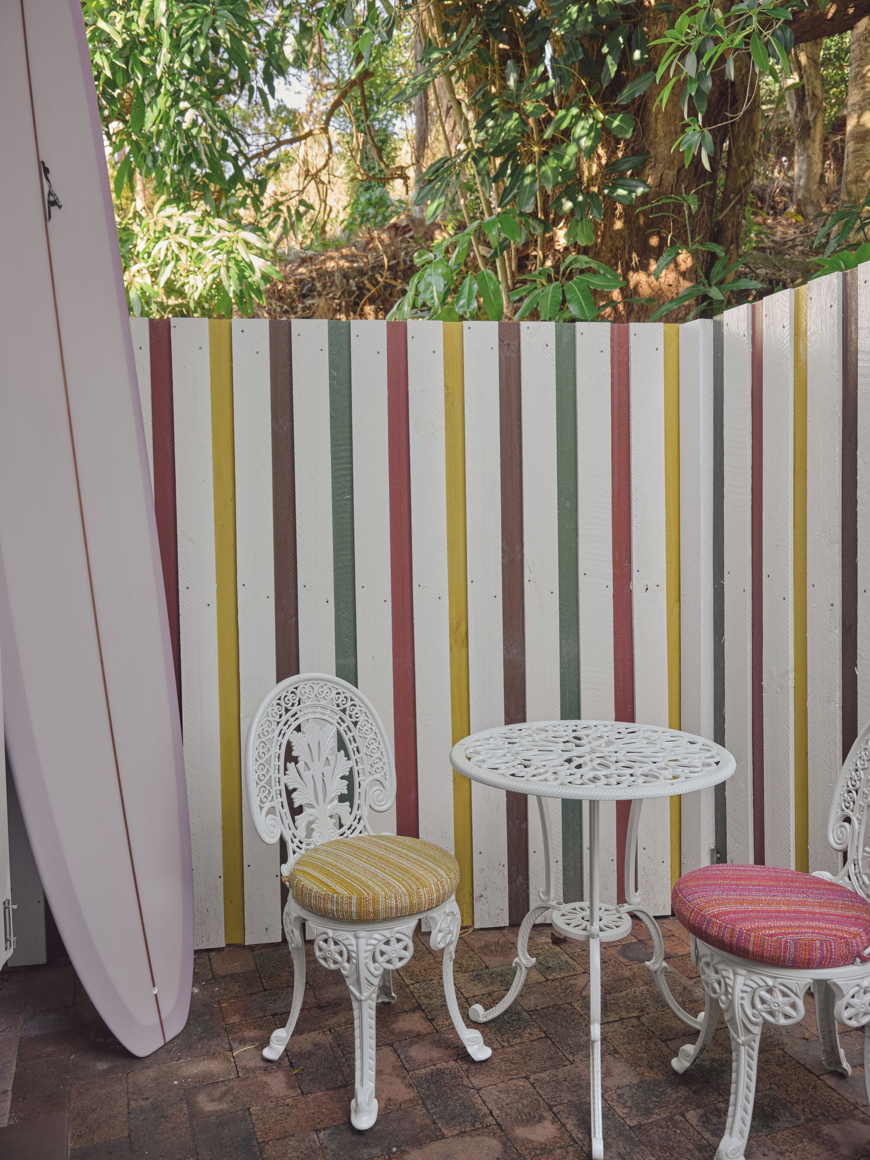 An outdoor seating area with two white metal chairs and a small round table, set against a striped fence.