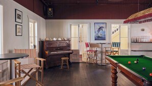 Une pièce équipée d’une table de billard, d’un piano et d’une table de jeu de fléchettes. On y trouve également des chaises et une petite table avec une lampe.