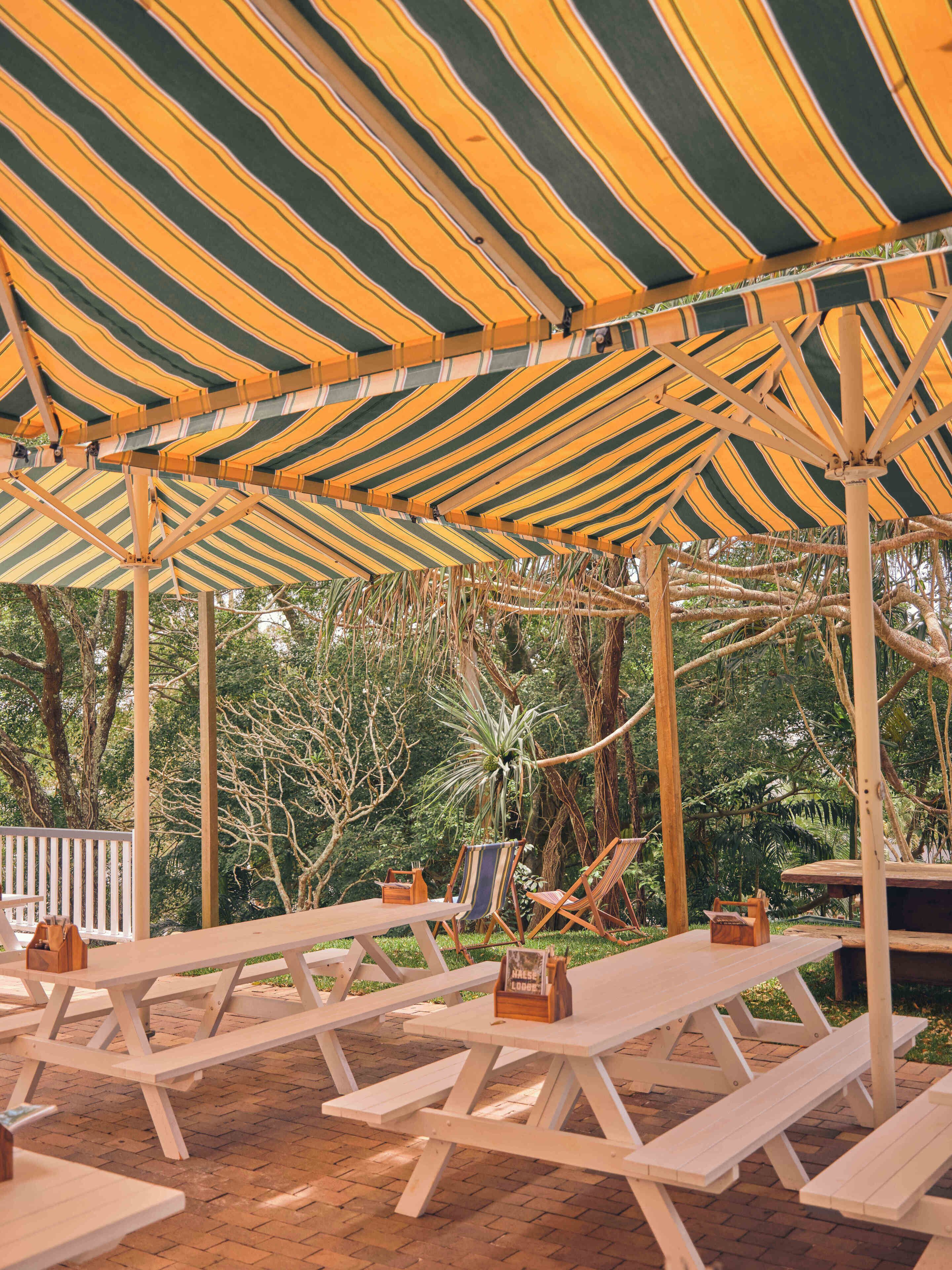 A covered outdoor seating area with picnic tables and chairs, surrounded by trees.