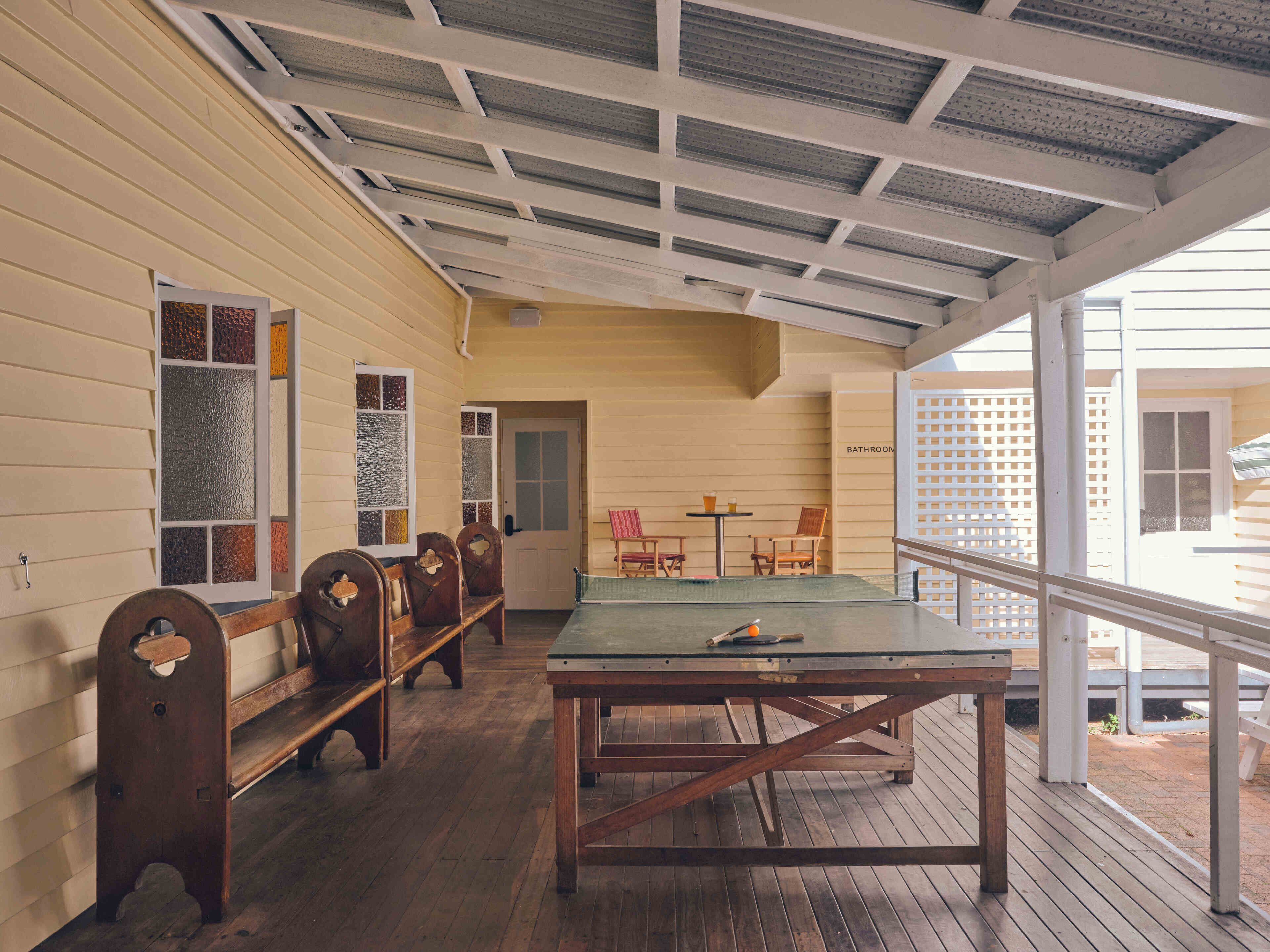 A covered porch with wooden benches and a ping pong table, a door leading to a bathroom, and a view of the outside.