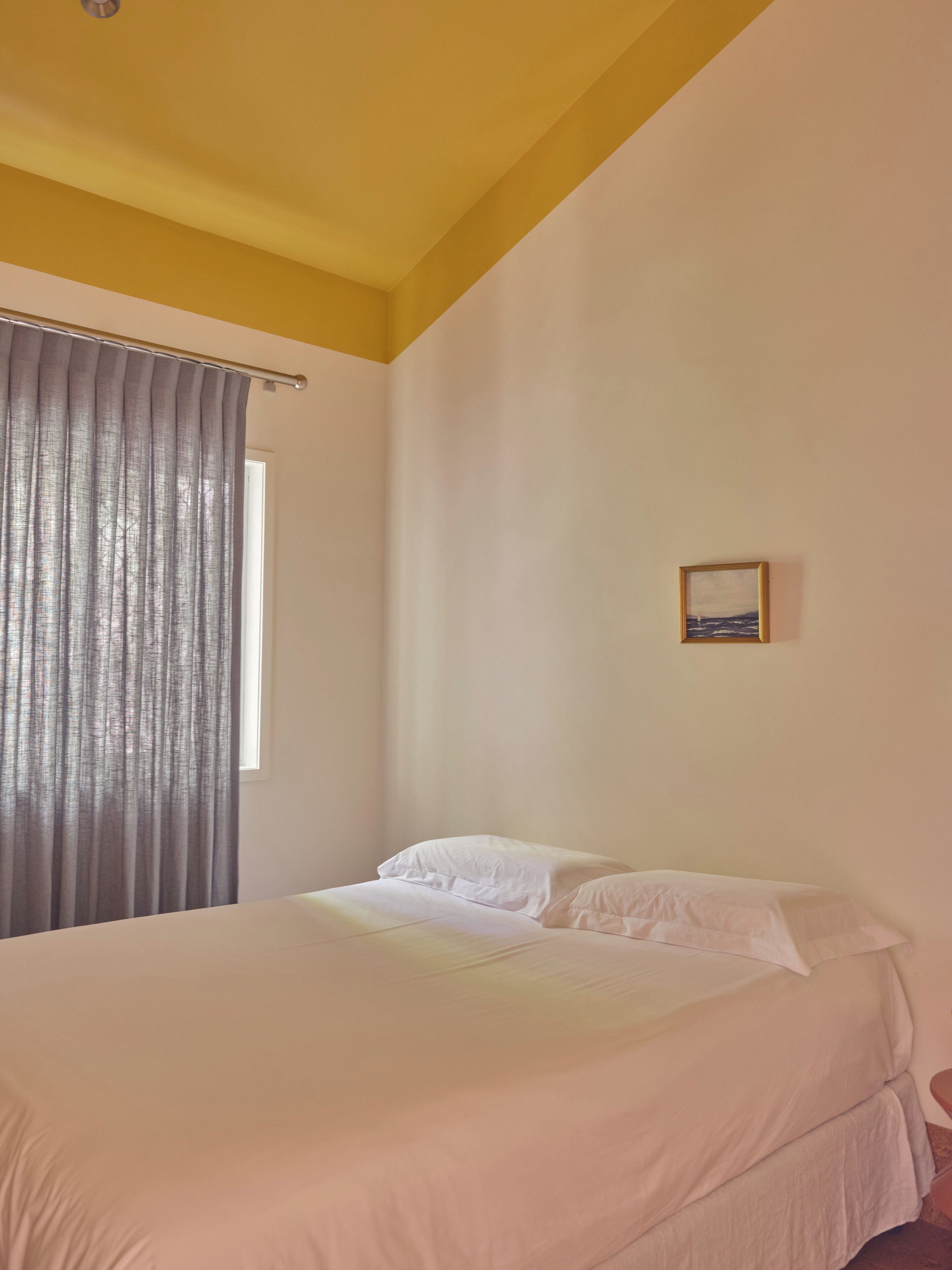 Un dormitorio con cama, ventana con cortinas y un pequeño cuadro en la pared.