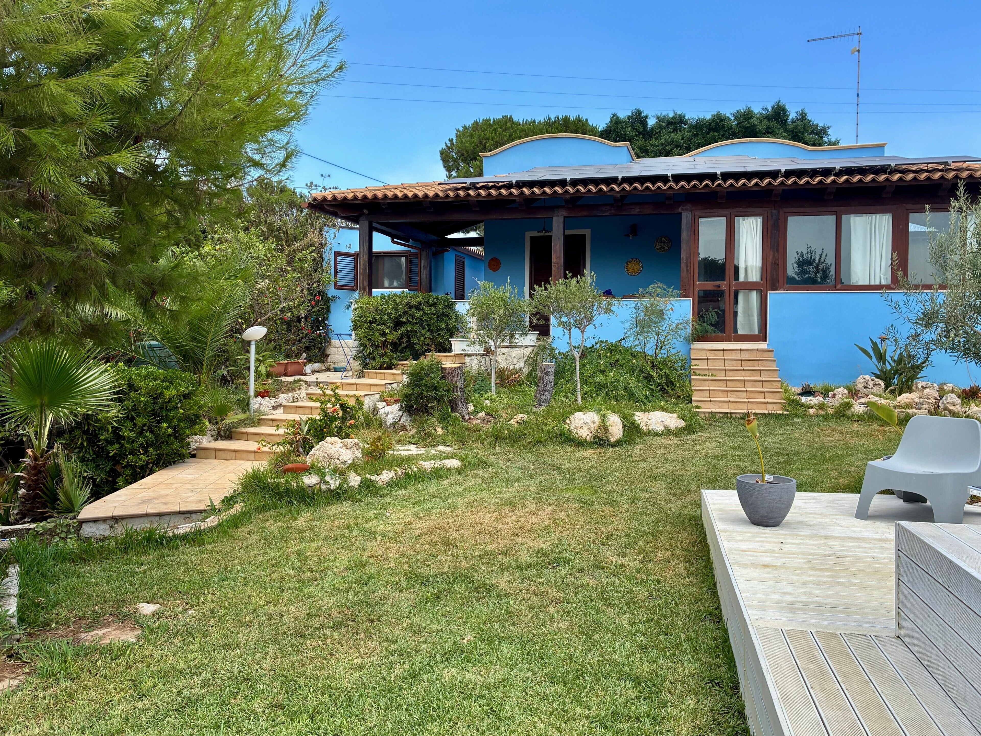 Uma casa com fachada azul, um deck de madeira e um jardim com diversas plantas e um pequeno lance de escadas que leva à entrada.