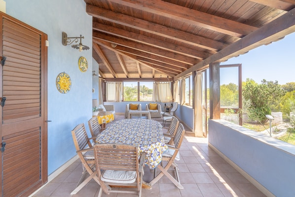 An outdoor patio with a wooden table and chairs, a covered area with a view of greenery, and a blue wall with decorative plates.