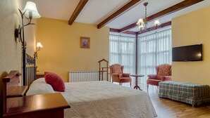 Quarto de hotel com cama, duas poltronas, uma mesa pequena, televisão e janela com cortinas.