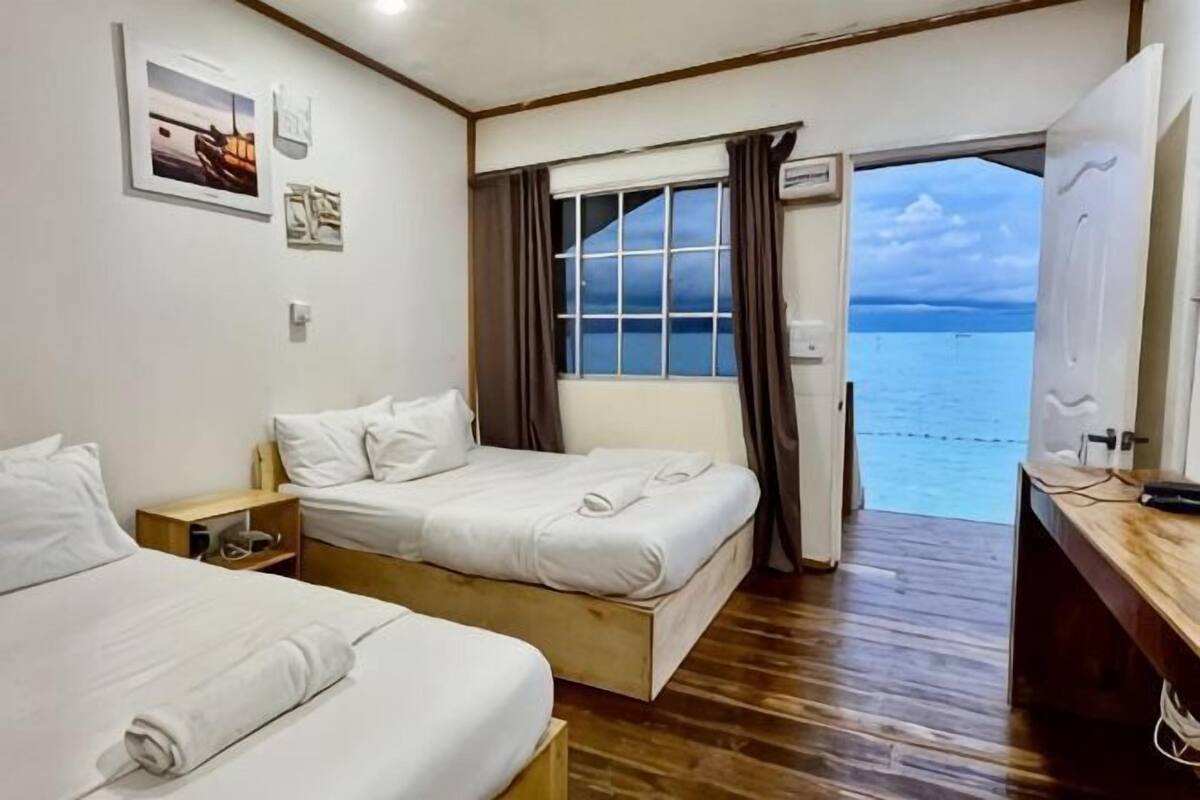 Une chambre avec deux lits, une fenêtre avec des rideaux, une porte donnant sur l’extérieur et une vue sur la mer.