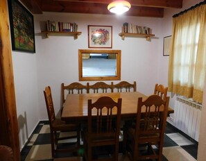 Uma sala de jantar com mesa e cadeiras de madeira, um espelho, estantes e uma janela com cortinas.