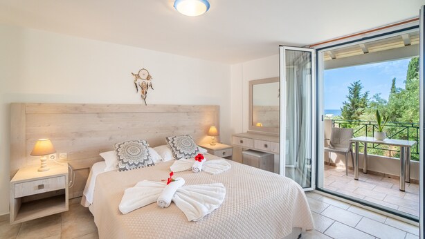 Ein Hotelzimmer mit einem großen Bett, zwei Nachttischen mit Lampen, einem kleinen Tisch und einem Balkon mit Stuhl und Tisch, mit Blick auf Grünflächen.