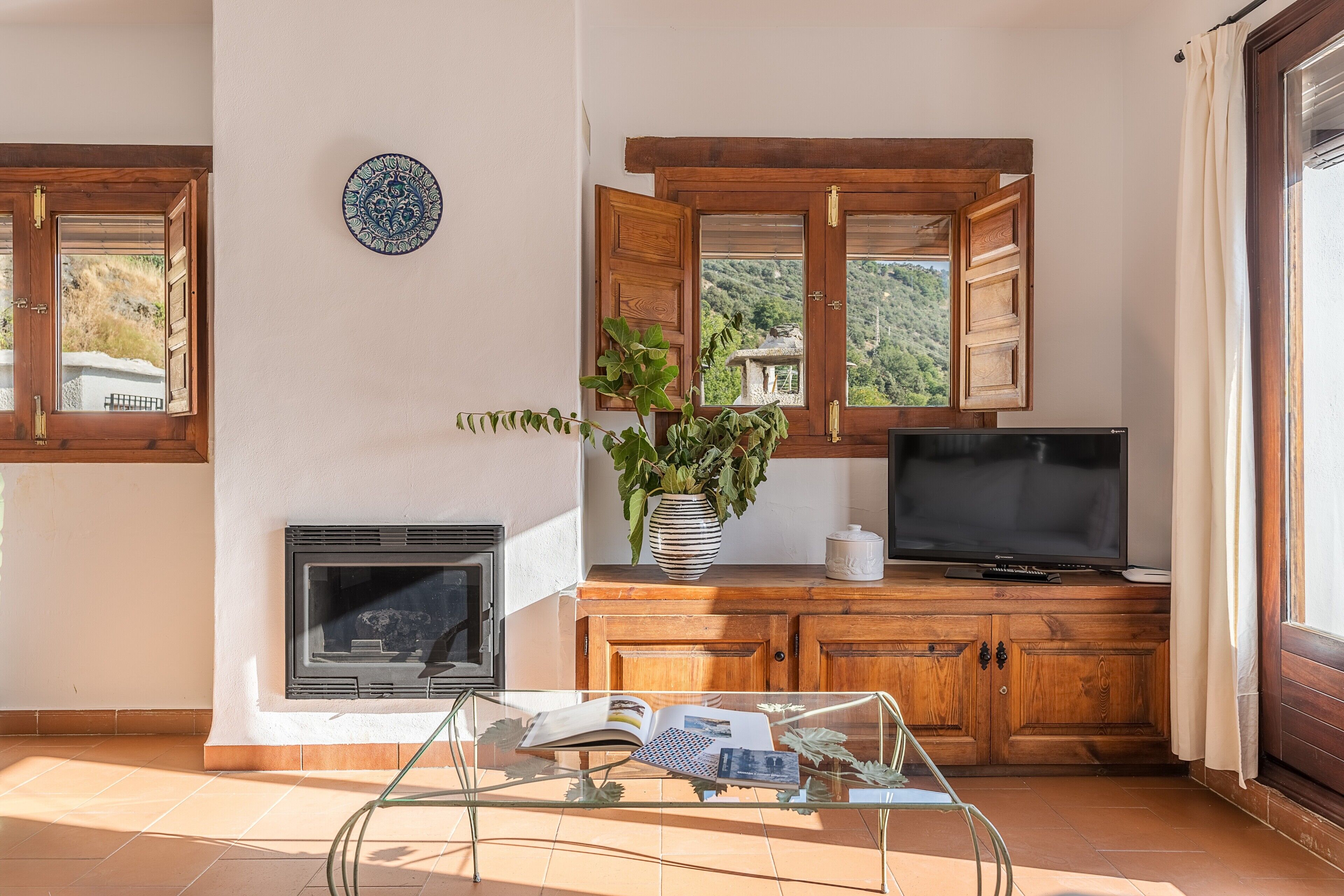 A living room with a fireplace, a television, a wooden cabinet, and a window with a view of a mountainous landscape.