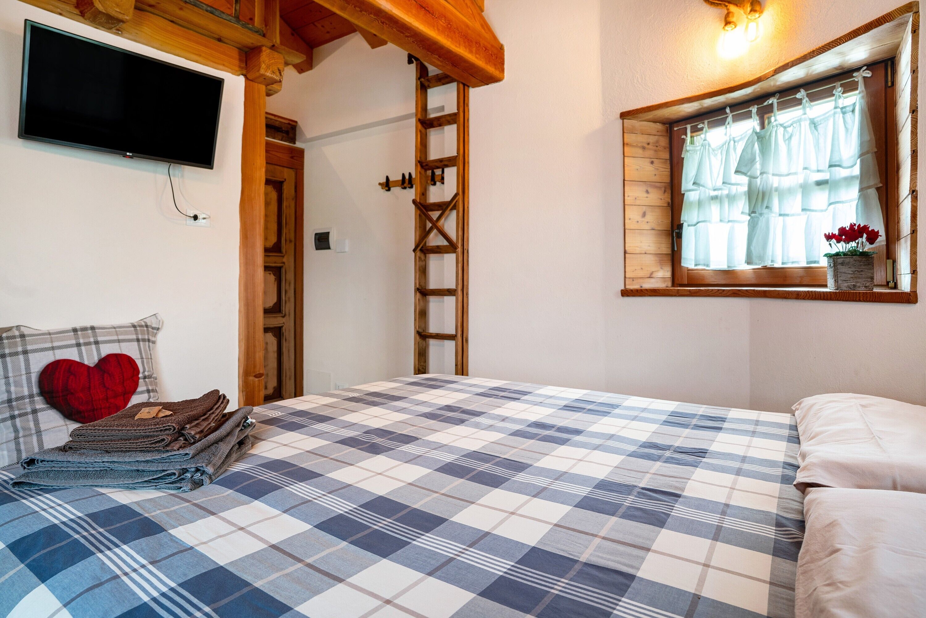 Une chambre avec une couvre-lit à carreaux, un coussin en forme de cœur, un téléviseur fixé au mur, une échelle en bois et une fenêtre avec des rideaux.