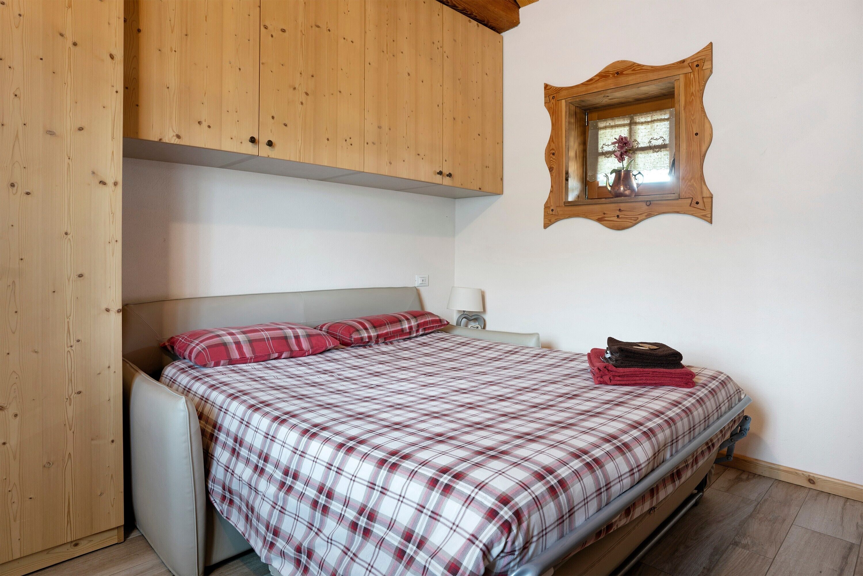 A bedroom with a bed, a wooden wardrobe, a mirror, and a window.