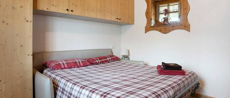 A bedroom with a bed, a wooden wardrobe, a mirror, and a window.