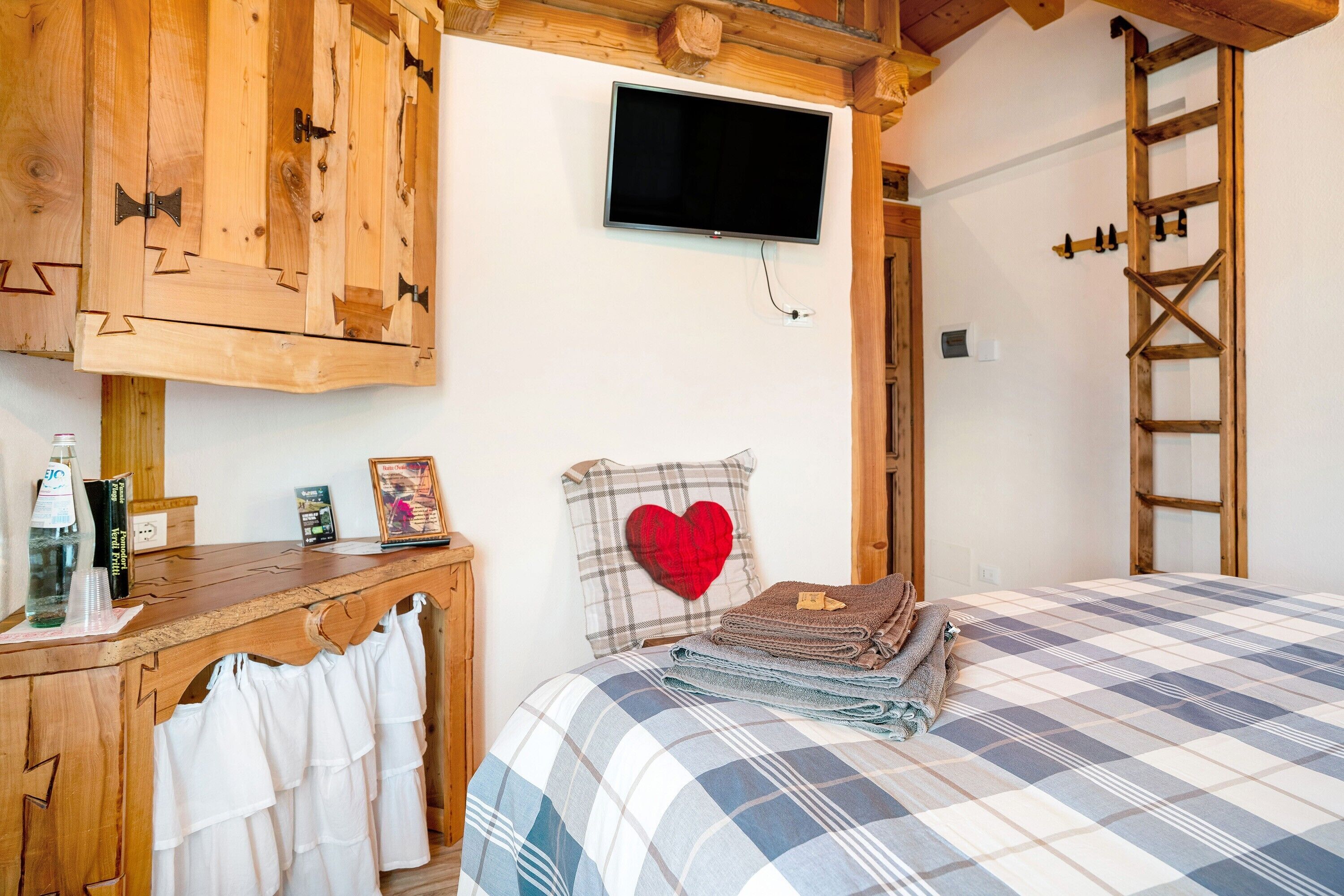 Una camera da letto con una coperta a quadri, una TV a muro, mobili in legno, una scrivania con una bottiglia e un cuscino a forma di cuore.