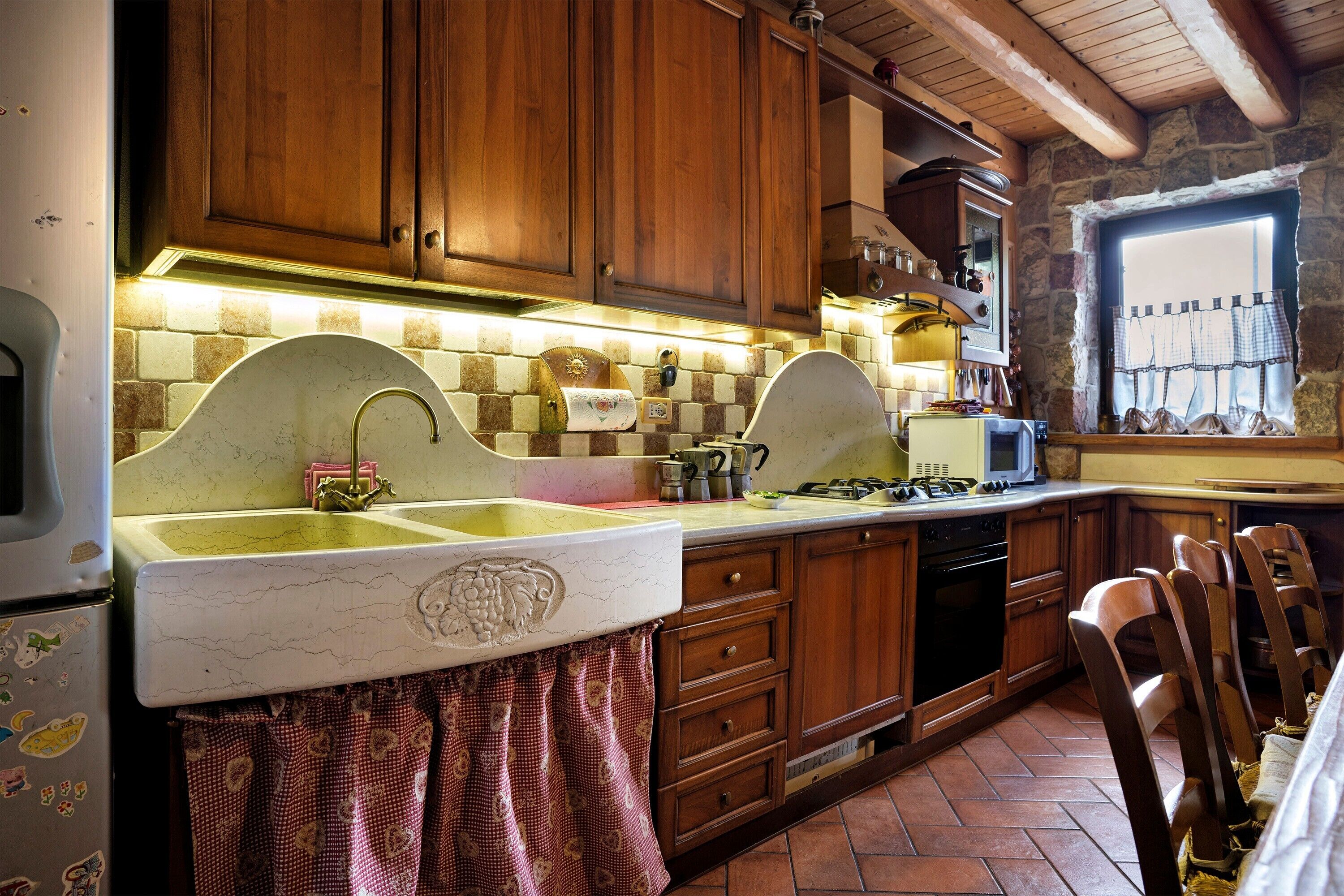 A kitchen with wooden cabinets, a marble countertop, and a window with curtains.