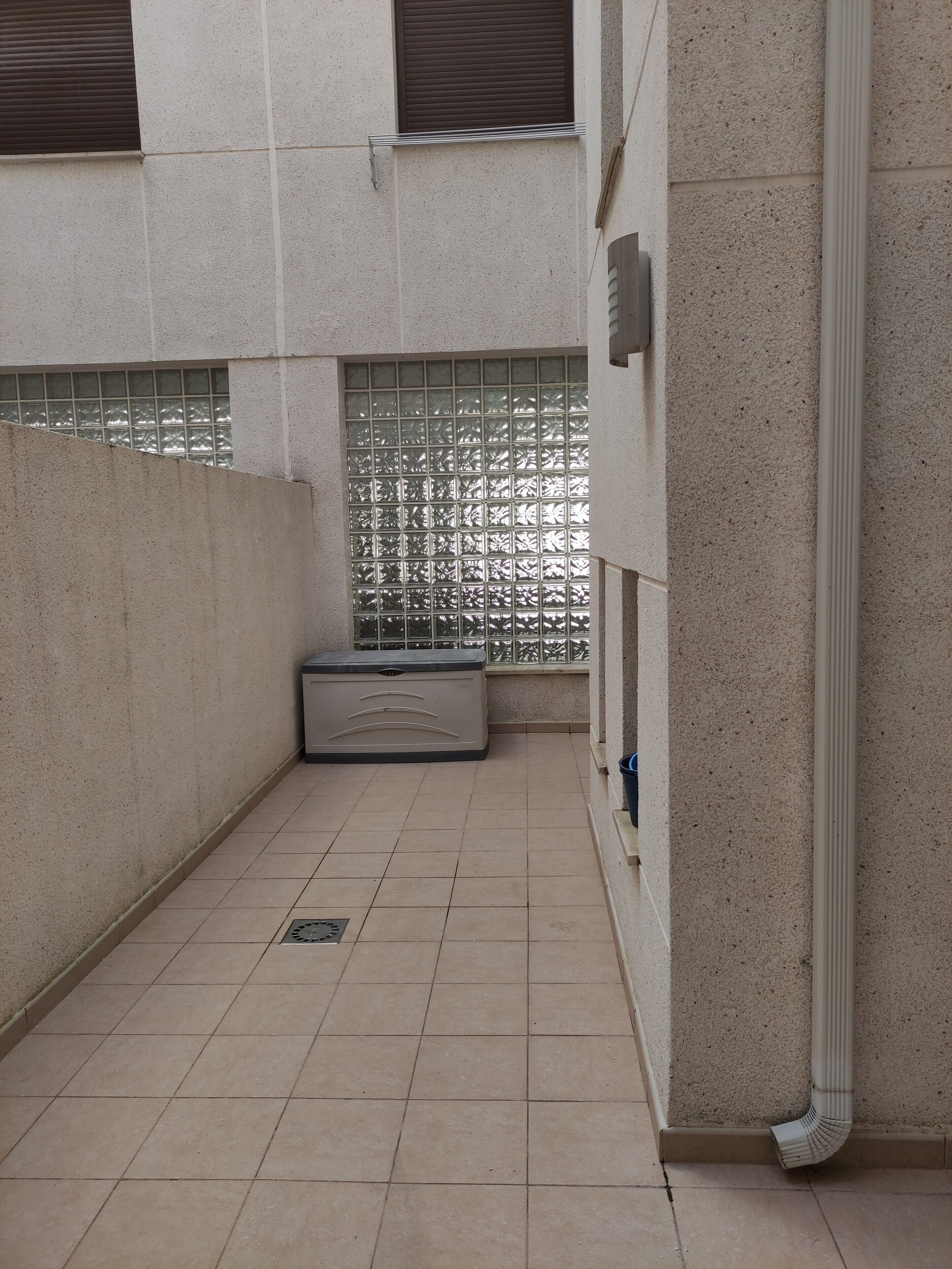 A narrow corridor with tiled flooring, a metal bench, a window with a metal grill, and a white pipe.