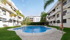 Una piscina rodeada por un área pavimentada, césped, palmeras y un edificio con balcones.
