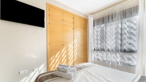 Una habitación de hotel con una cama, un televisor montado en la pared y un ventanal con cortinas ligeras.