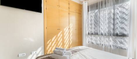 Una habitación de hotel con una cama, un televisor montado en la pared y un ventanal con cortinas ligeras.