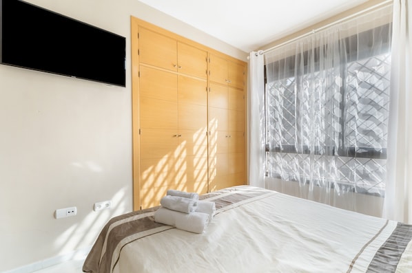 A hotel room with a bed, a TV mounted on the wall, and a large window with sheer curtains.