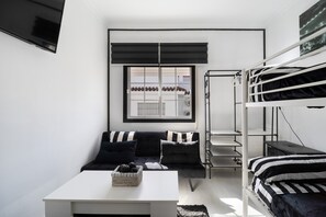 Ein modernes Zimmer mit einem Etagenbett, einer Couch mit gestreiften Kissen, einem weißen Tisch und einem Fenster mit Jalousien.