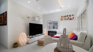 A modern living room with a flat-screen TV, white brick wall, white sofa, and a colorful abstract painting.