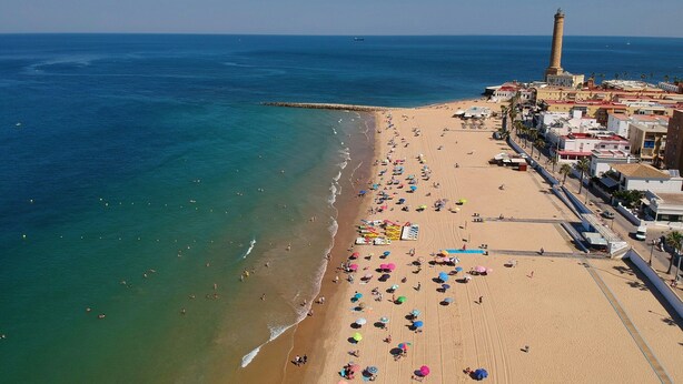 An aerial view of a beach with a lighthouse, beach umbrellas, and a promenade with buildings.