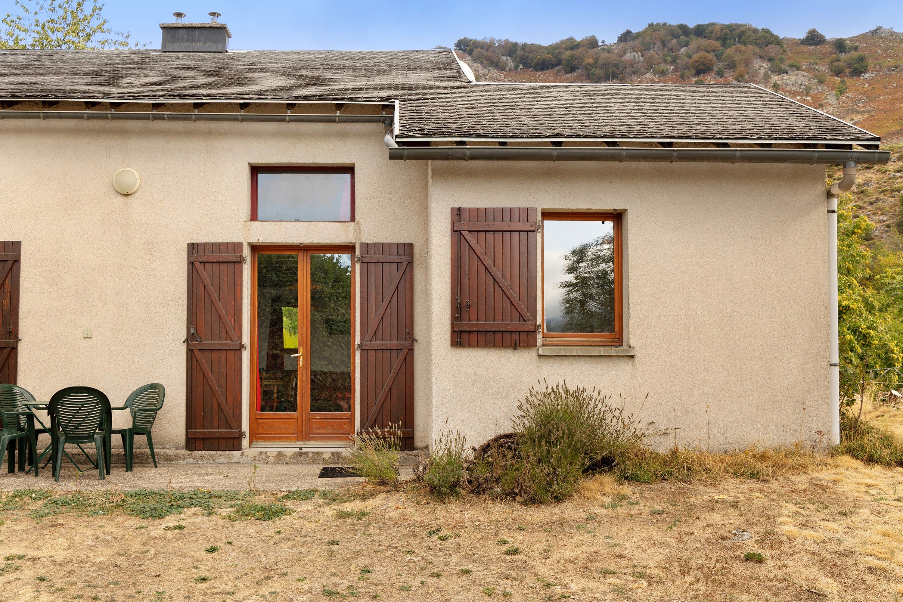 Un bâtiment à deux étages, doté d’un toit plat, de volets en bois aux fenêtres et d’un petit porche avec des chaises.