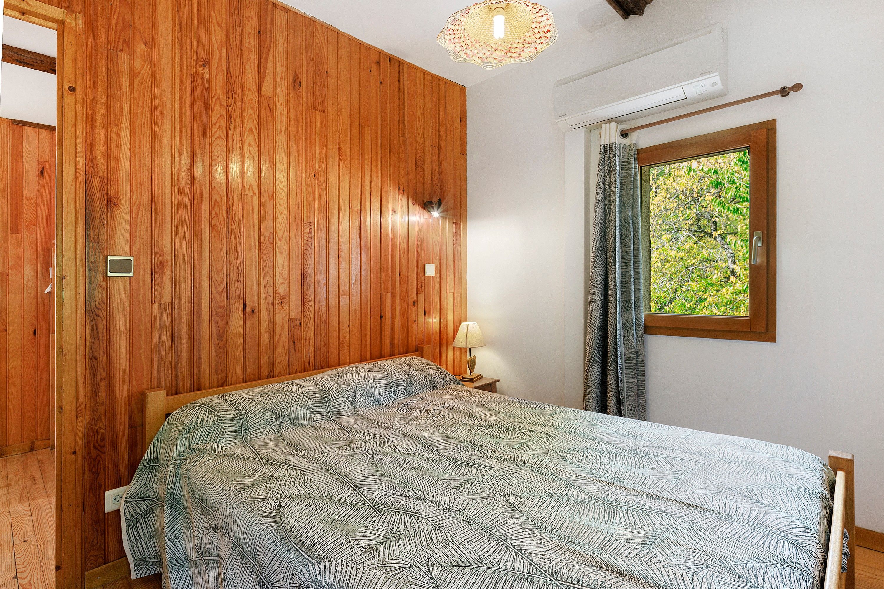 A bedroom with wooden panel walls, a bed with a patterned bedspread, a nightstand with a lamp, a window with curtains, and an air conditioning unit.