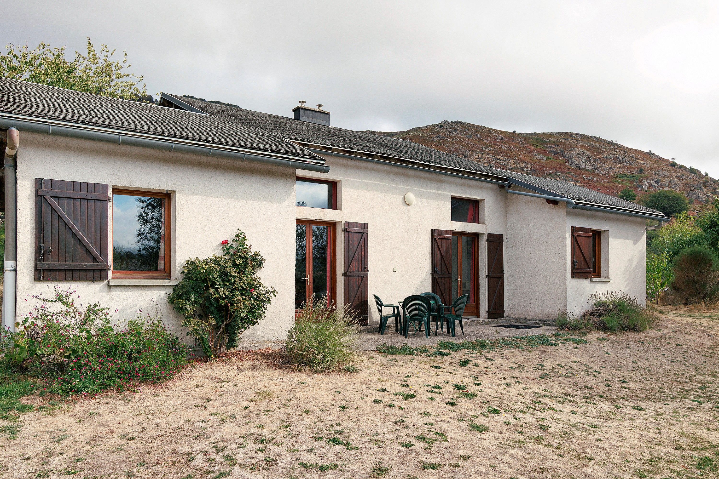 Ein zweigeschossiges Haus mit einem Ziegeldach, ein kleiner Garten mit Tisch und Stühlen sowie eine Hügelkulisse im Hintergrund.