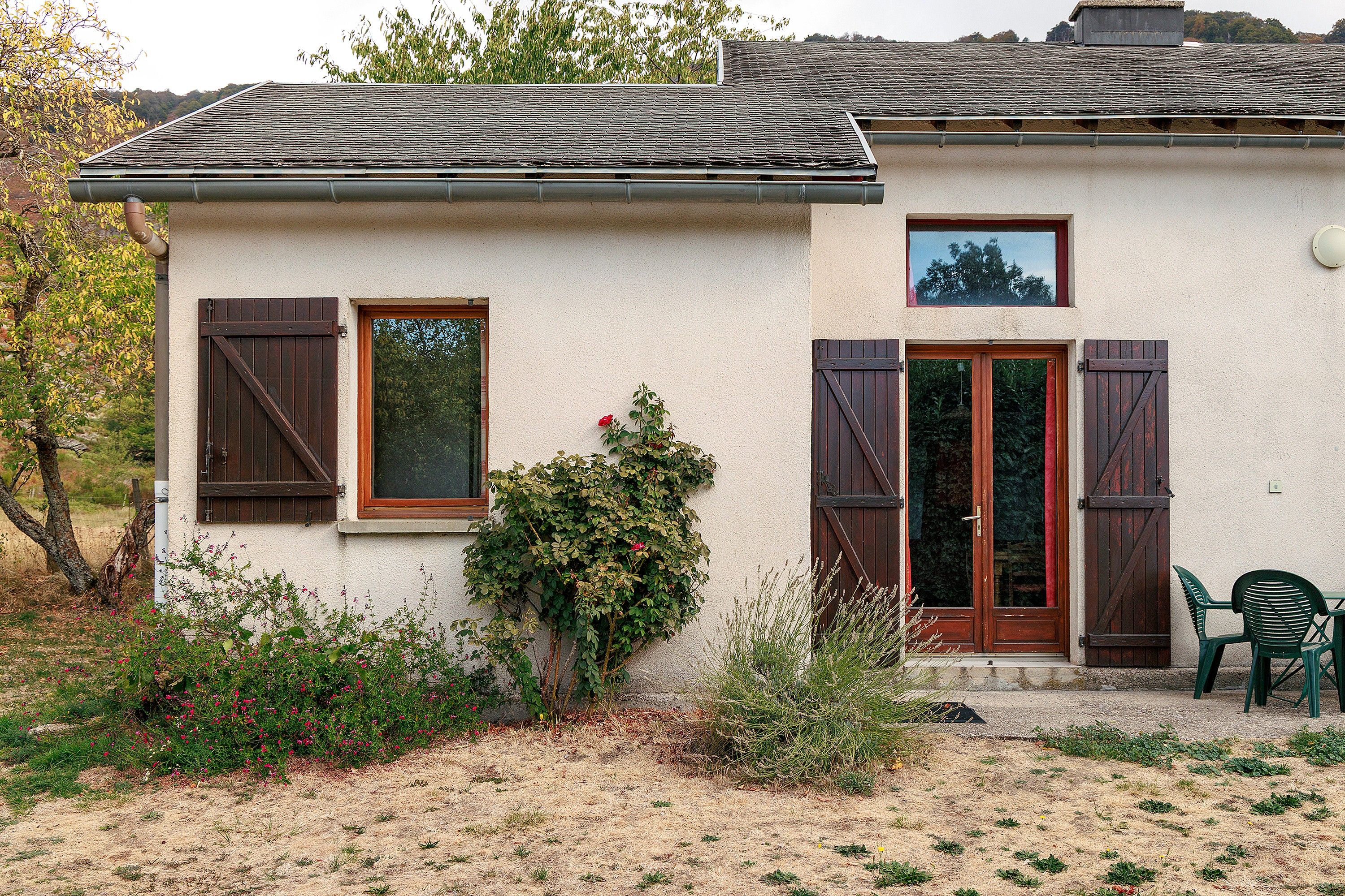 A rustic house with a tiled roof, wooden shutters, and a small garden with shrubs and flowers.