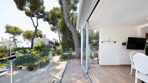 Un interior moderno con un gran ventanal que da a un jardín exterior, un televisor de pantalla plana en la pared y una silla blanca junto a la ventana.