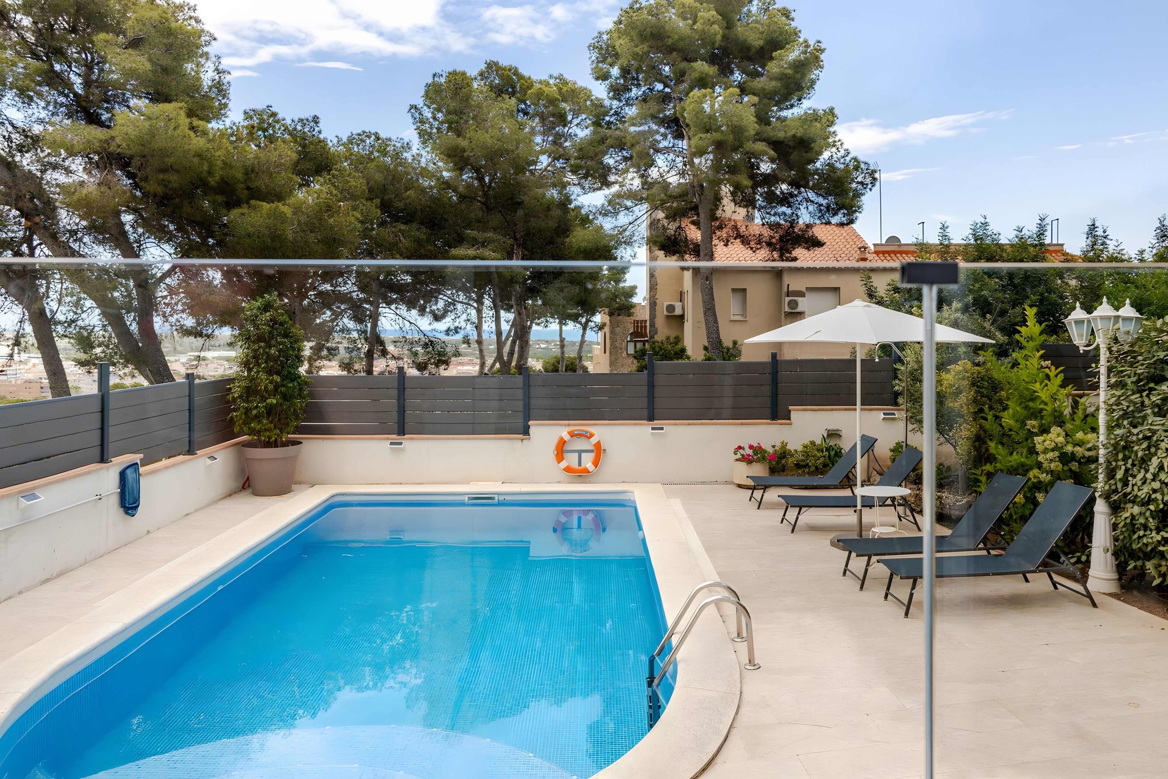 A swimming pool with a lifebuoy, lounge chairs, and a view of trees and buildings.