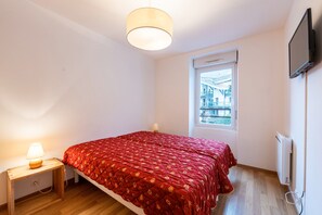 Un dormitorio con cama, lámpara de noche, ventana y televisor instalado en la pared.