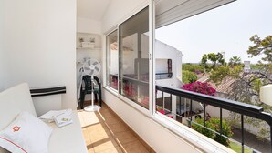 Un balcon avec un canapé blanc, un ventilateur et une vue sur un jardin et des immeubles.