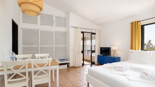 Une chambre d’hôtel avec un lit, un bureau avec des chaises, une télévision, une table de chevet bleue et une fenêtre avec des rideaux.