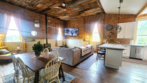 A rustic kitchen with wooden floors, a dining area with a wooden table and chairs, a sofa, and a television mounted on the wall. - Charming 3-bedroom historic Brick Loft in Uniontown with WiFi (Uniontown)