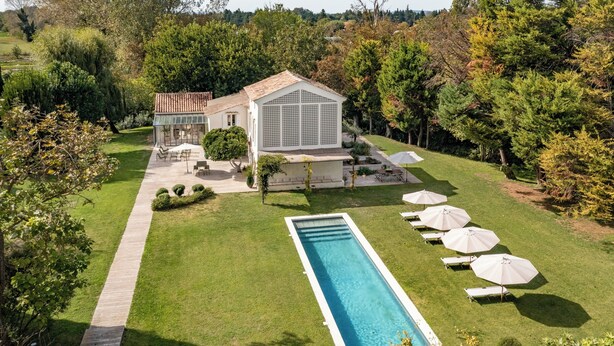 Una amplia piscina rectangular rodeada de césped cuidado, un patio con muebles de exterior y varias sombrillas.
