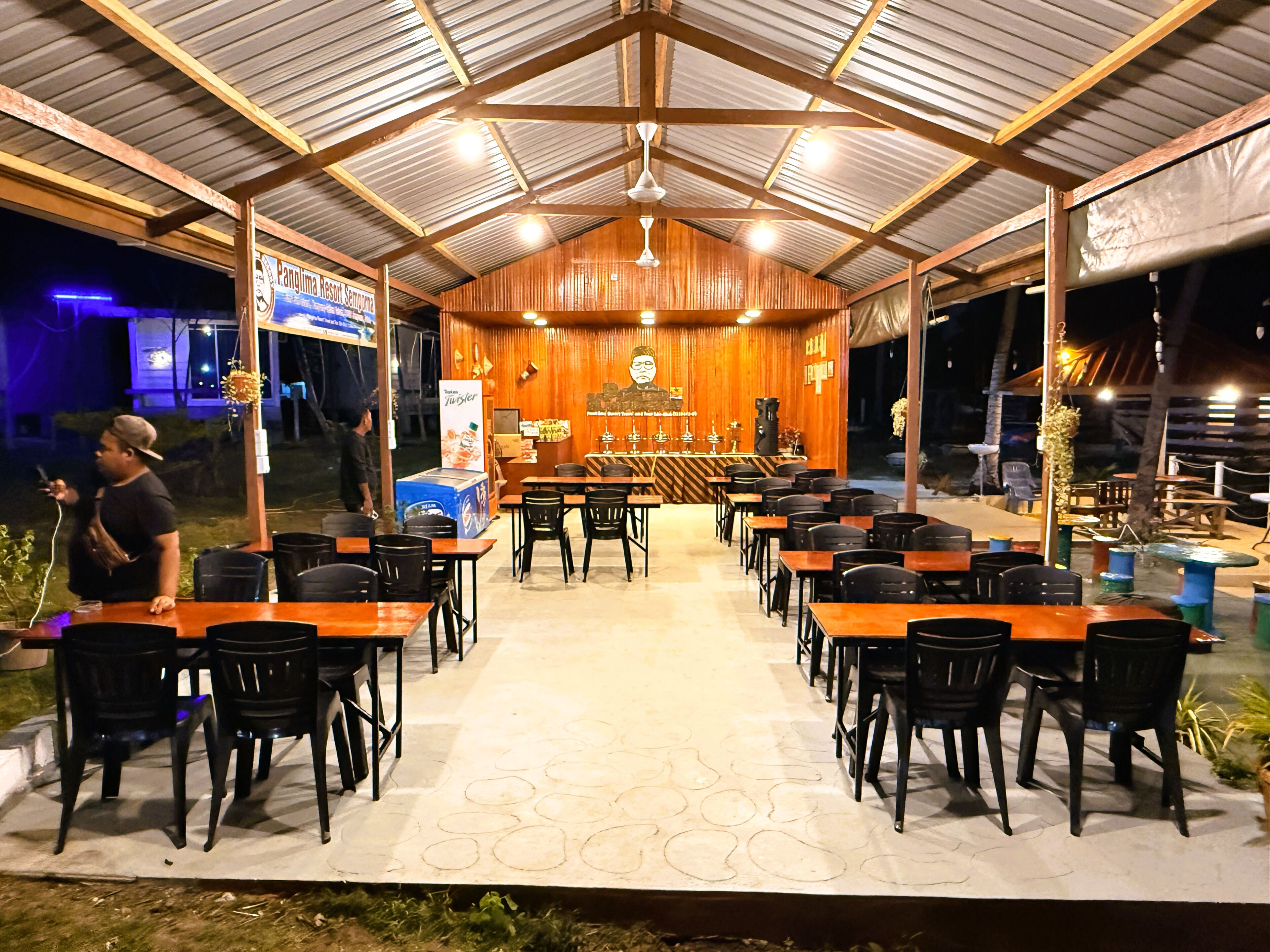 A covered outdoor dining area with tables and chairs, a bar in the center, and a wooden wall with a mural.
