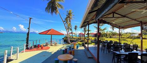 Un restaurant en bord de mer avec une terrasse, des palmiers et une vue sur l’océan.