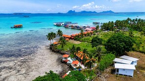 Vista aérea de un complejo turístico costero con bungalows sobre el agua, una playa y montañas al fondo.