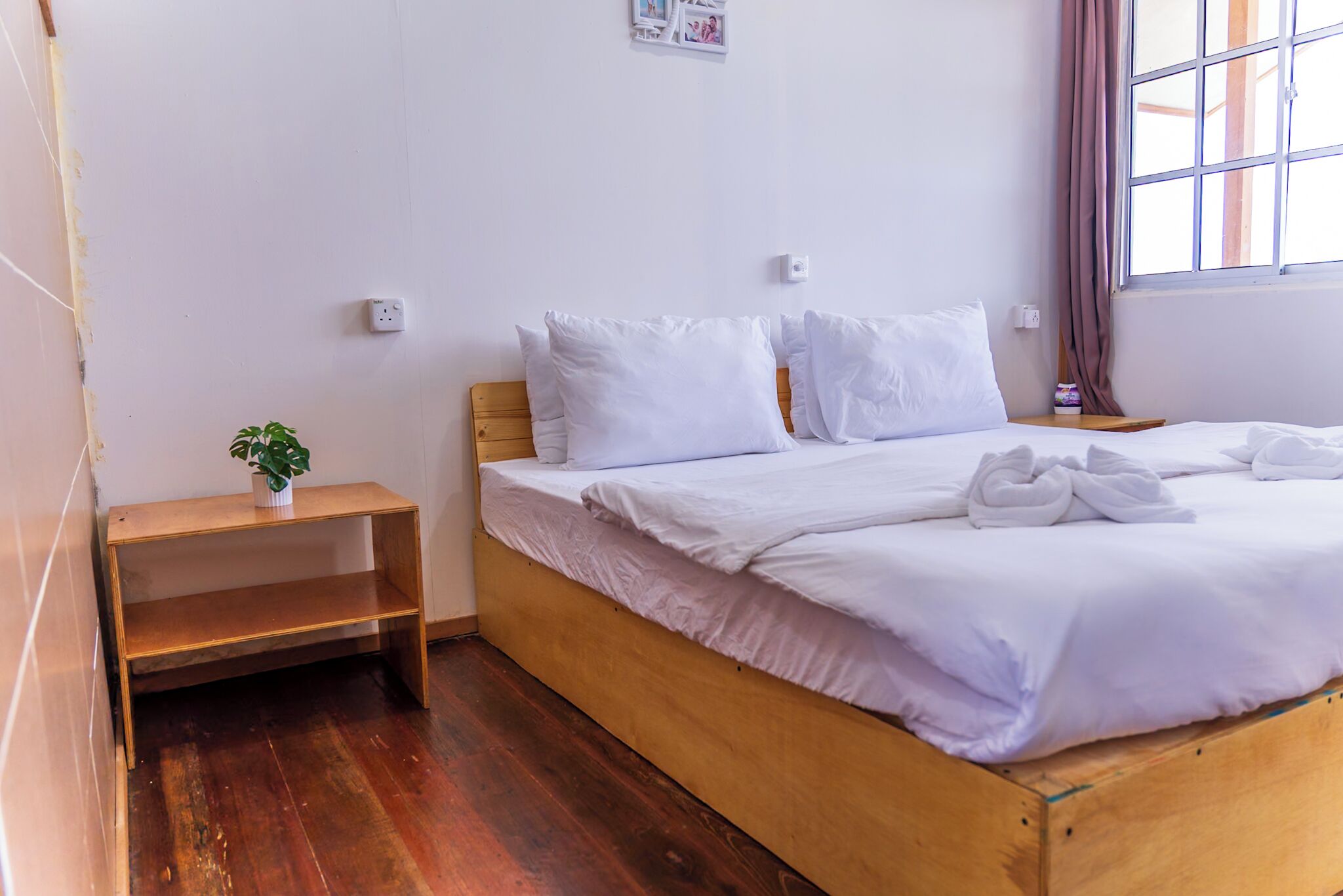 Una cama bien hecha con ropa de cama y toallas blancas, una mesita de noche de madera con una planta y una ventana con cortinas.