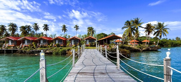 Un muelle de madera que conduce a bungalós sobre el agua con techos rojos, rodeado de palmeras y agua turquesa cristalina.