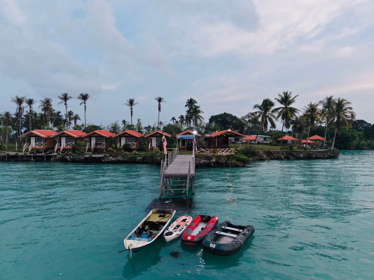A dock with several boats, a pier leading to a row of overwater bungalows with red roofs, and palm trees in the background.